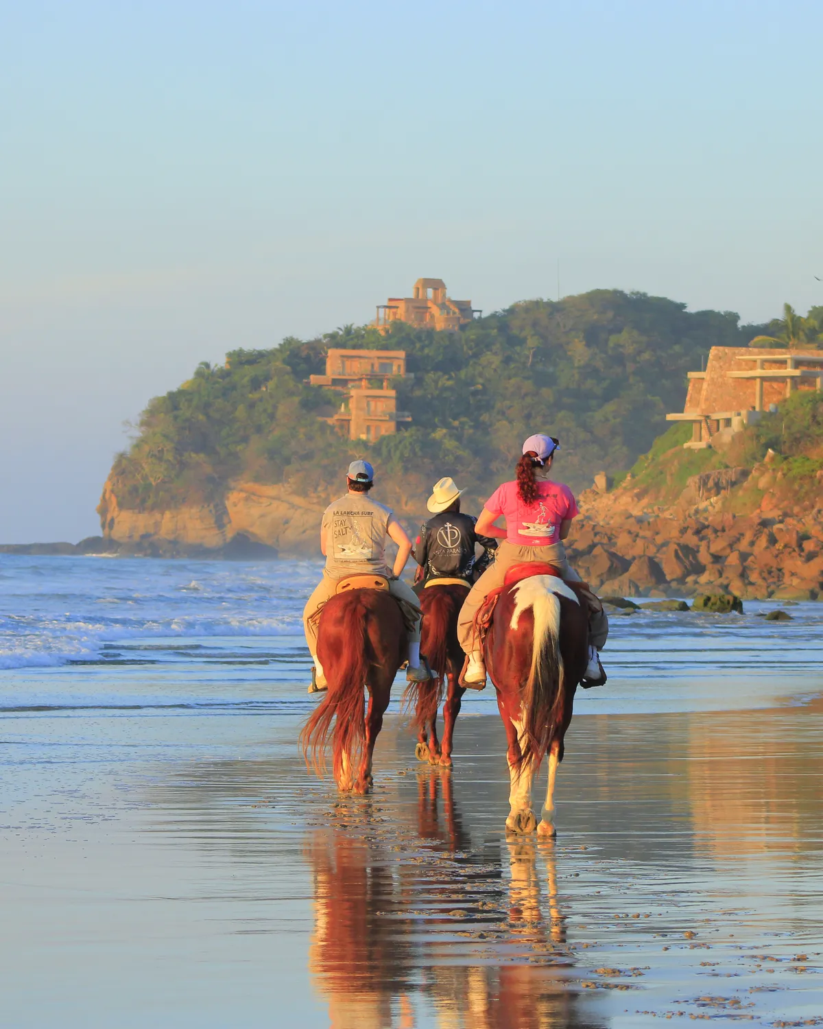 Riders on horseback down the wet sand at golden hour, cliff villas on the point behind