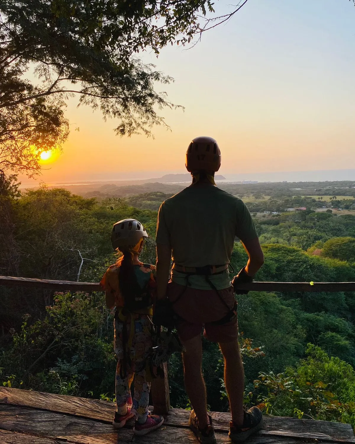A father and child in zipline helmets at the sunset overlook, the valley running down to the sea