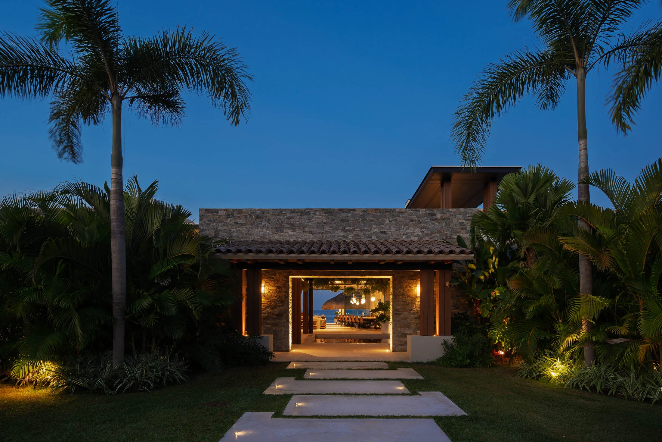 The stone entry at dusk — the lit path leading to a doorway that frames the palapa and the Pacific
