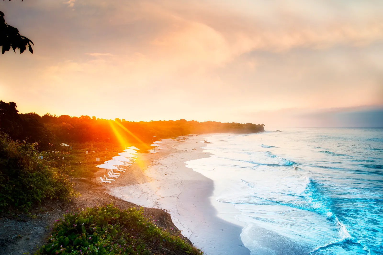 The coastline at El Surf Club at golden hour — sun flare on the water and surf running down the point