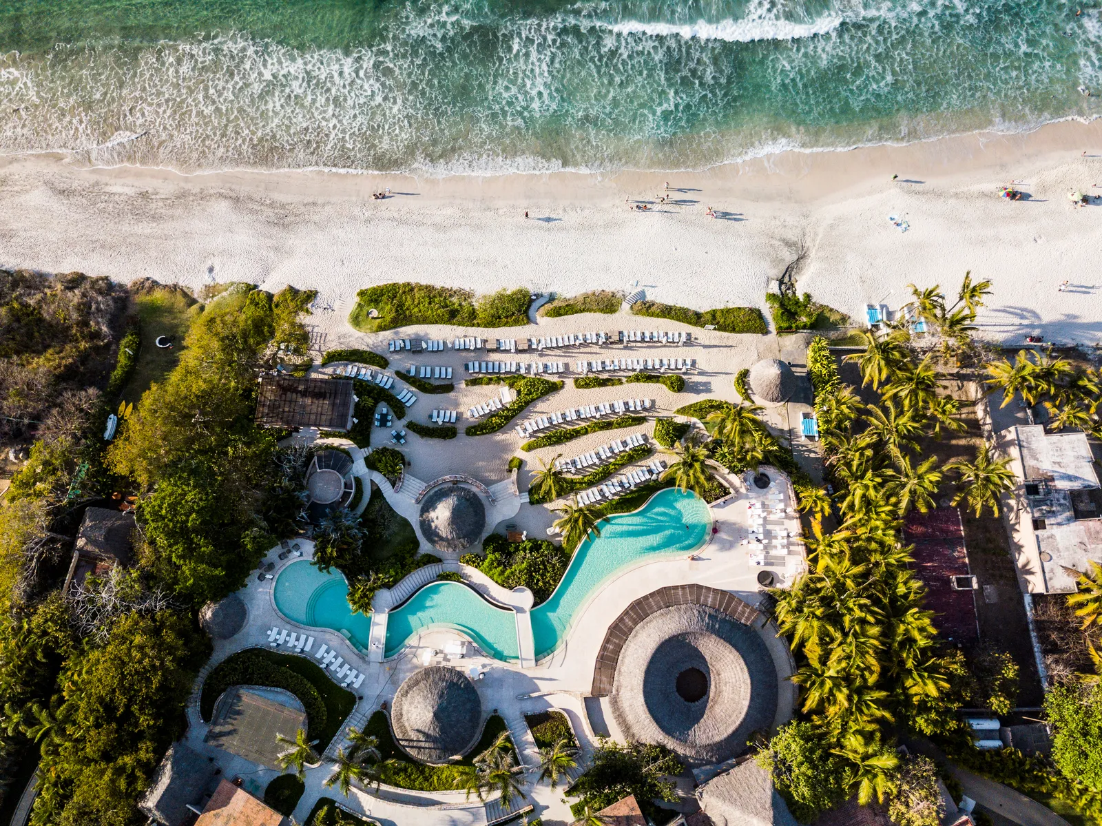 Kupuri Beach Club from directly above — the pool winding through loungers to the wide sand and surf of Litibú Bay