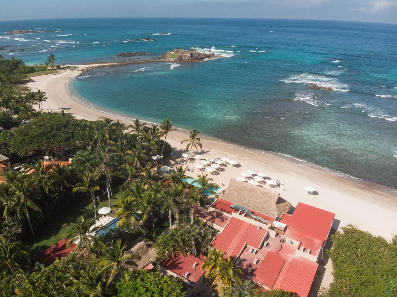 Pacífico Beach Club from above — red-tile roofs and palms on a white-sand cove, reef water beyond