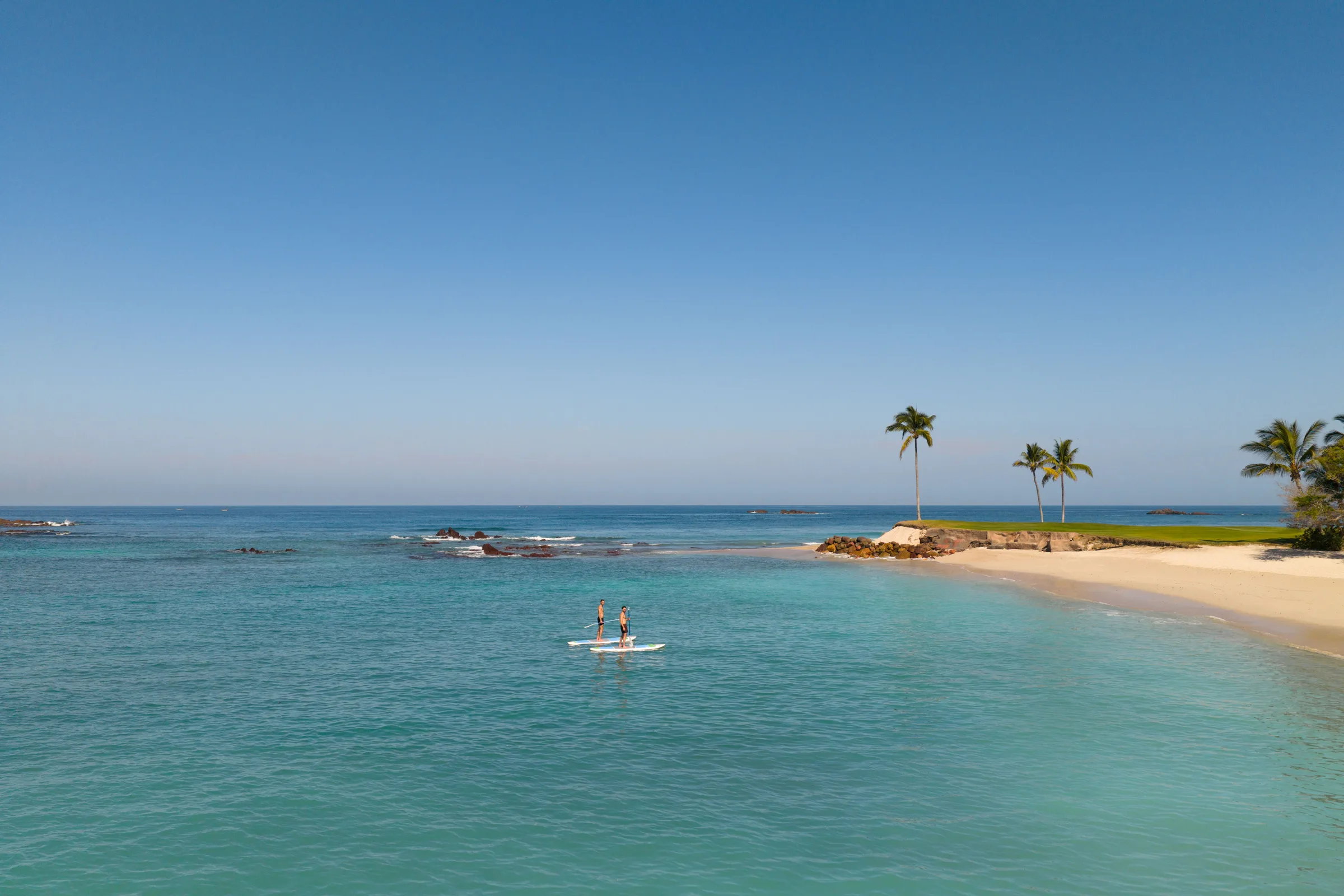 Two paddleboarders crossing the calm turquoise cove at Sea Breeze, white sand and palms on the point beyond