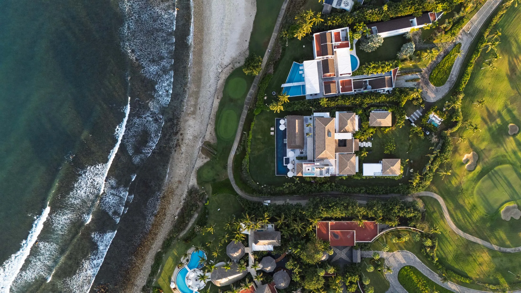 A top-down aerial of the estate between the beach and the fairways — the pool, the palapa and the lawns from directly above