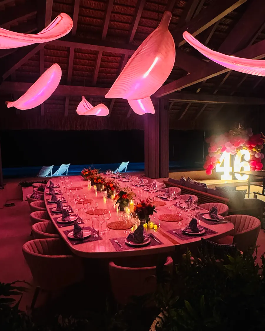 A birthday dinner under the palapa — the table glowing pink beneath lit palm sculptures, marquee numbers by the pool