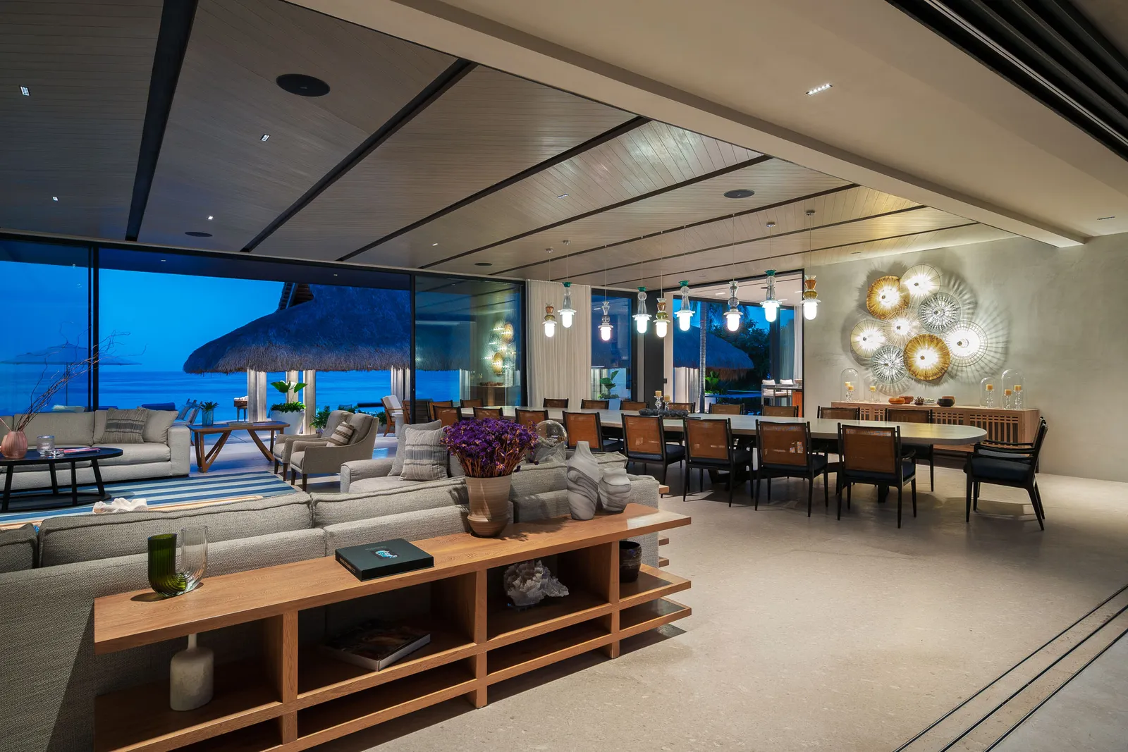 The great room at blue hour — the sofa field, the long dining table and the palapa beyond the glass