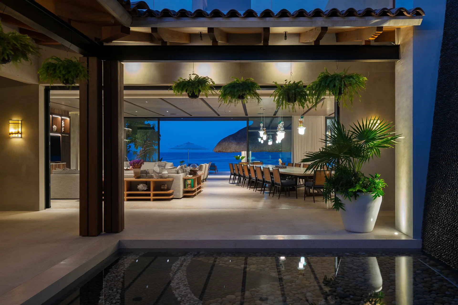 Looking through the great room from the entry court — ferns overhead, sofas and the long table, the ocean beyond