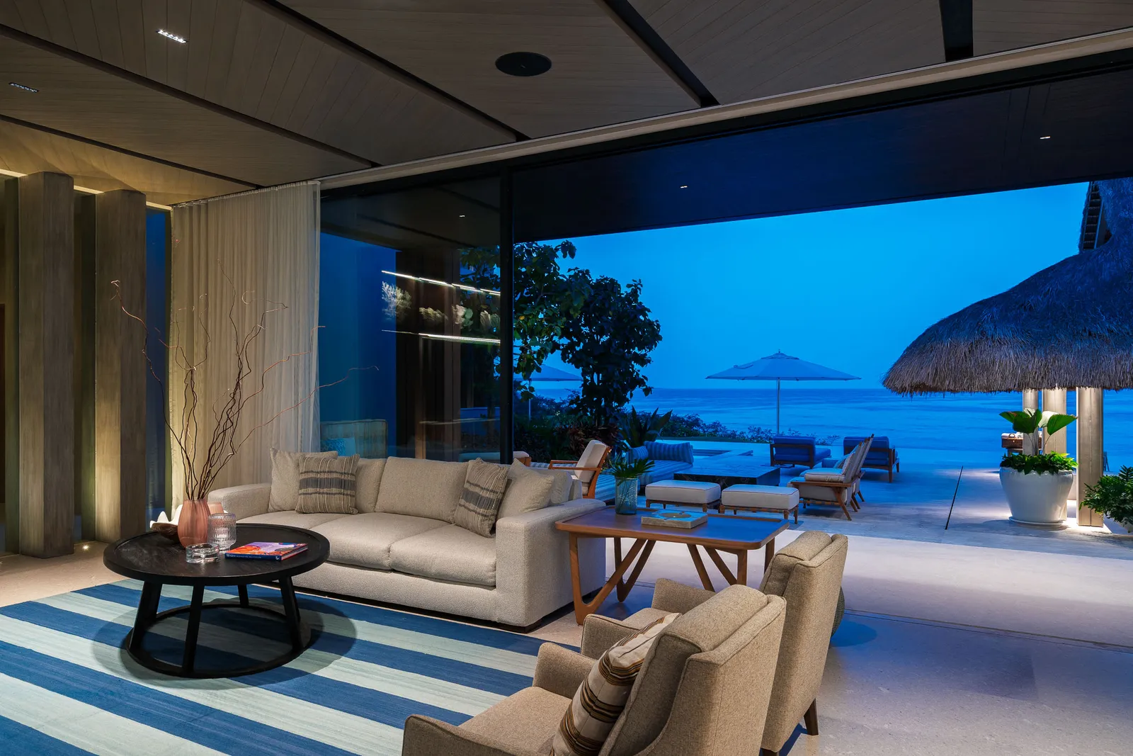 The lounge corner at dusk, doors slid away to the terrace, umbrella and ocean in the blue light