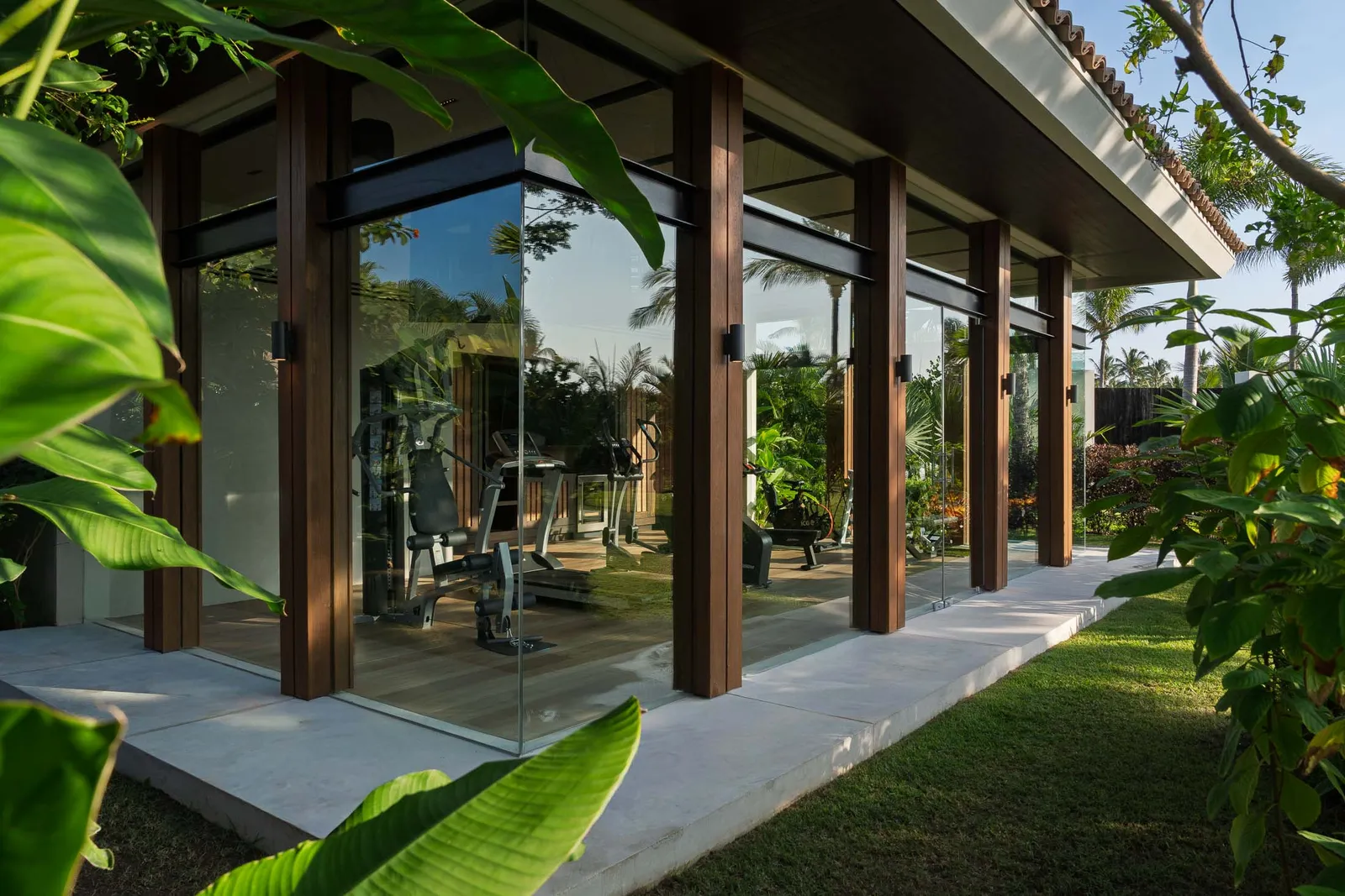 The fitness studio from the arrival lawn — a glass pavilion of its own, machines lit behind full-height glass among the leaves