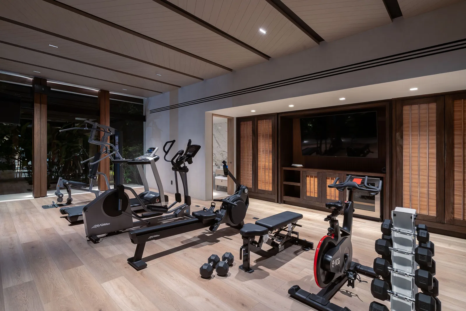 Inside the fitness studio in the evening — cardio row, dumbbells and the screen wall, the garden dark beyond the glass