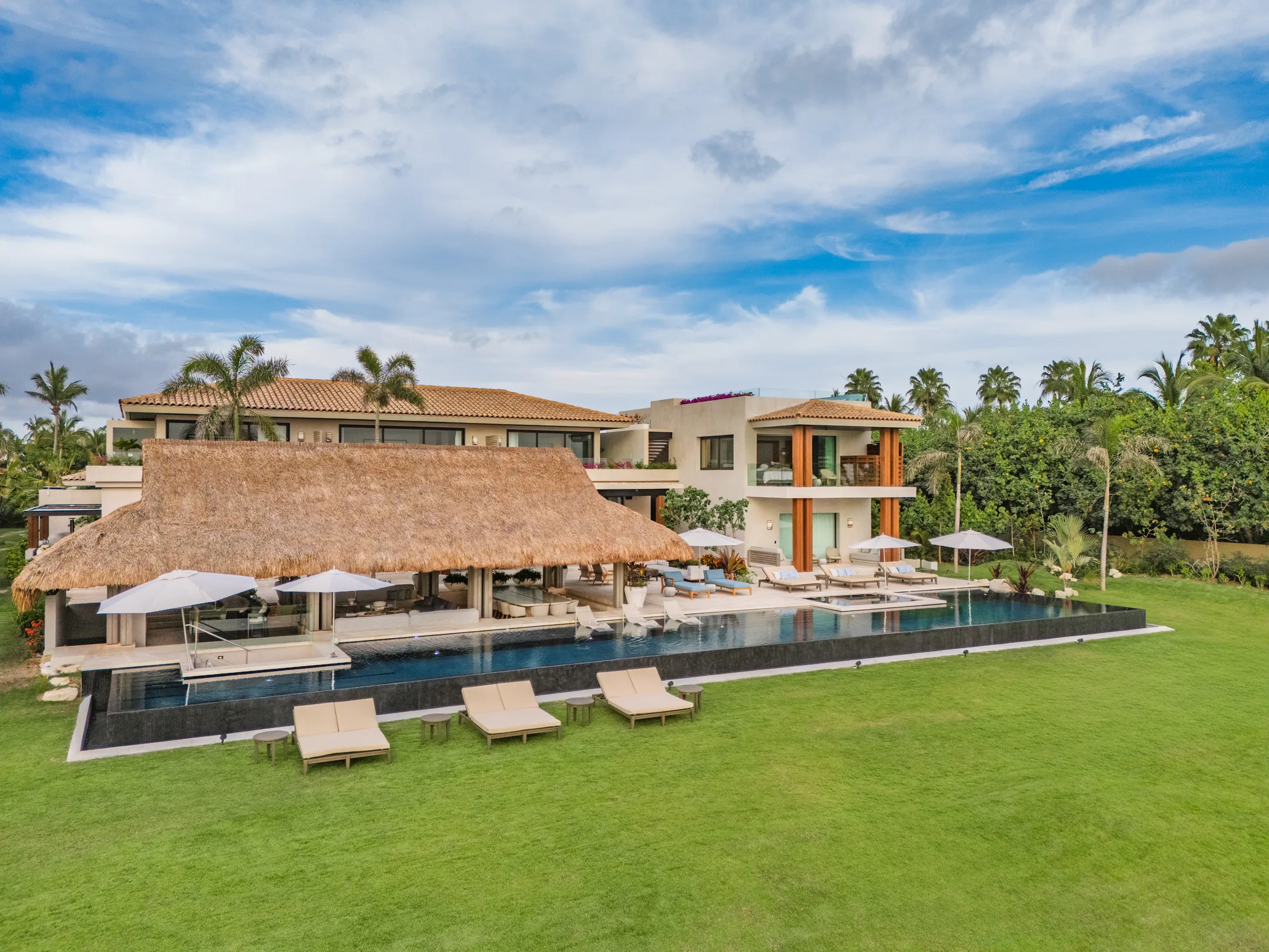 The whole estate head-on from above the lawn — the palapa, the infinity pool and both wings under a midday sky
