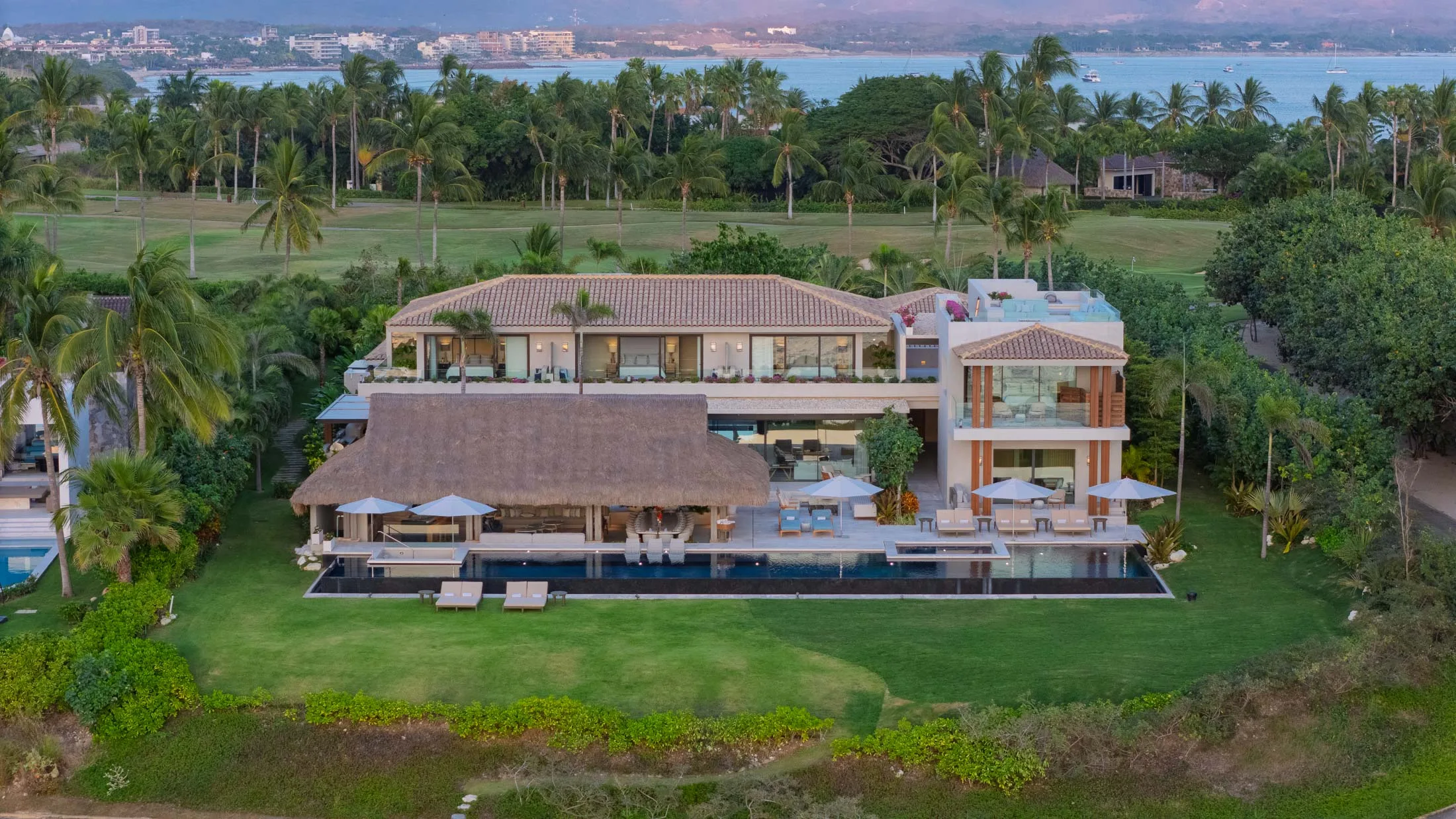 Casa de Elementos at dusk from above — the palapa, the lit suites and the infinity pool between the lawn and the fairways