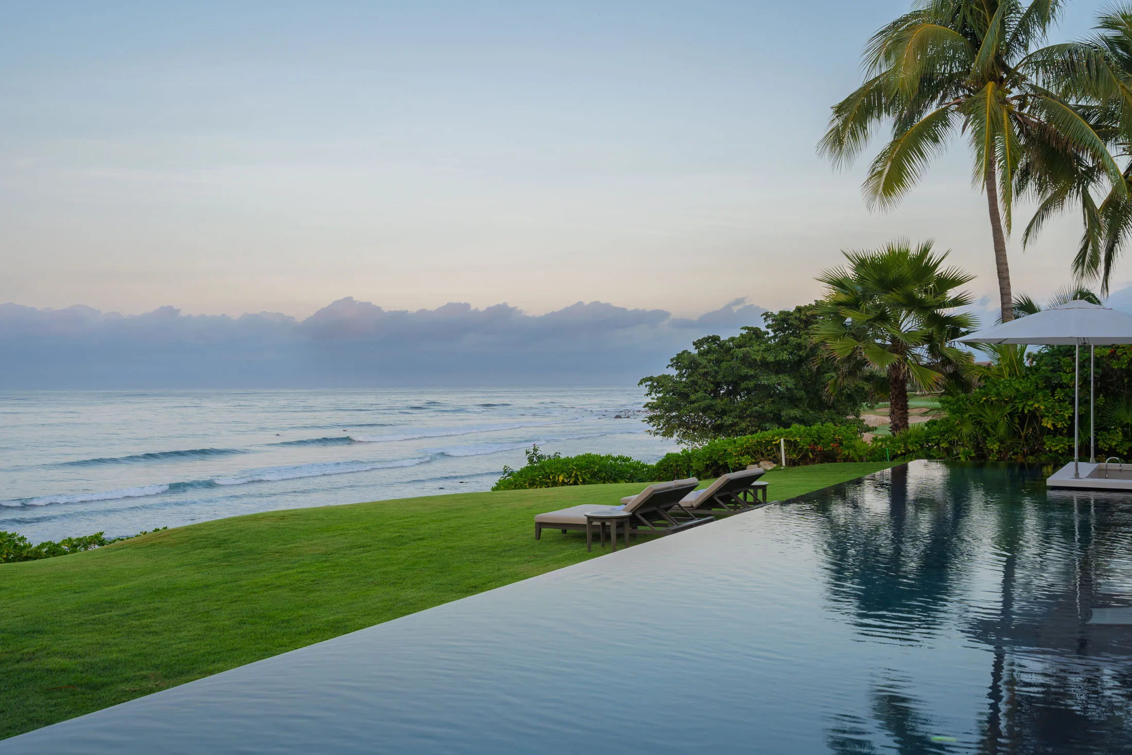 The infinity pool at dawn, glass-calm — loungers on the lawn, waves rolling in beyond the edge