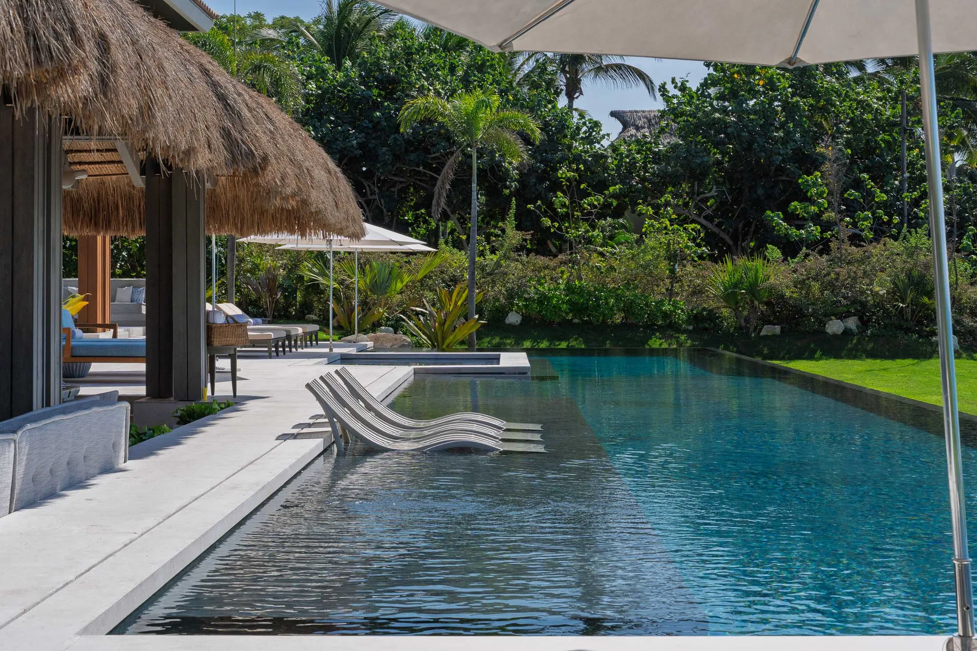 The pool at midday — in-water loungers on the shelf, the lane running past the palapa