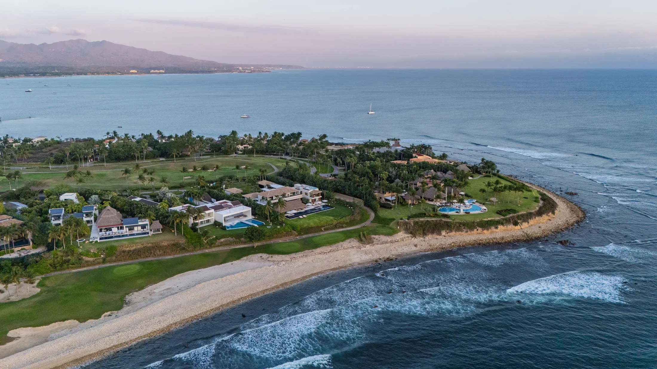 La Punta at golden hour from above — Casa de Elementos and the point’s villas between the fairways and the surf, Banderas Bay and the mountains beyond