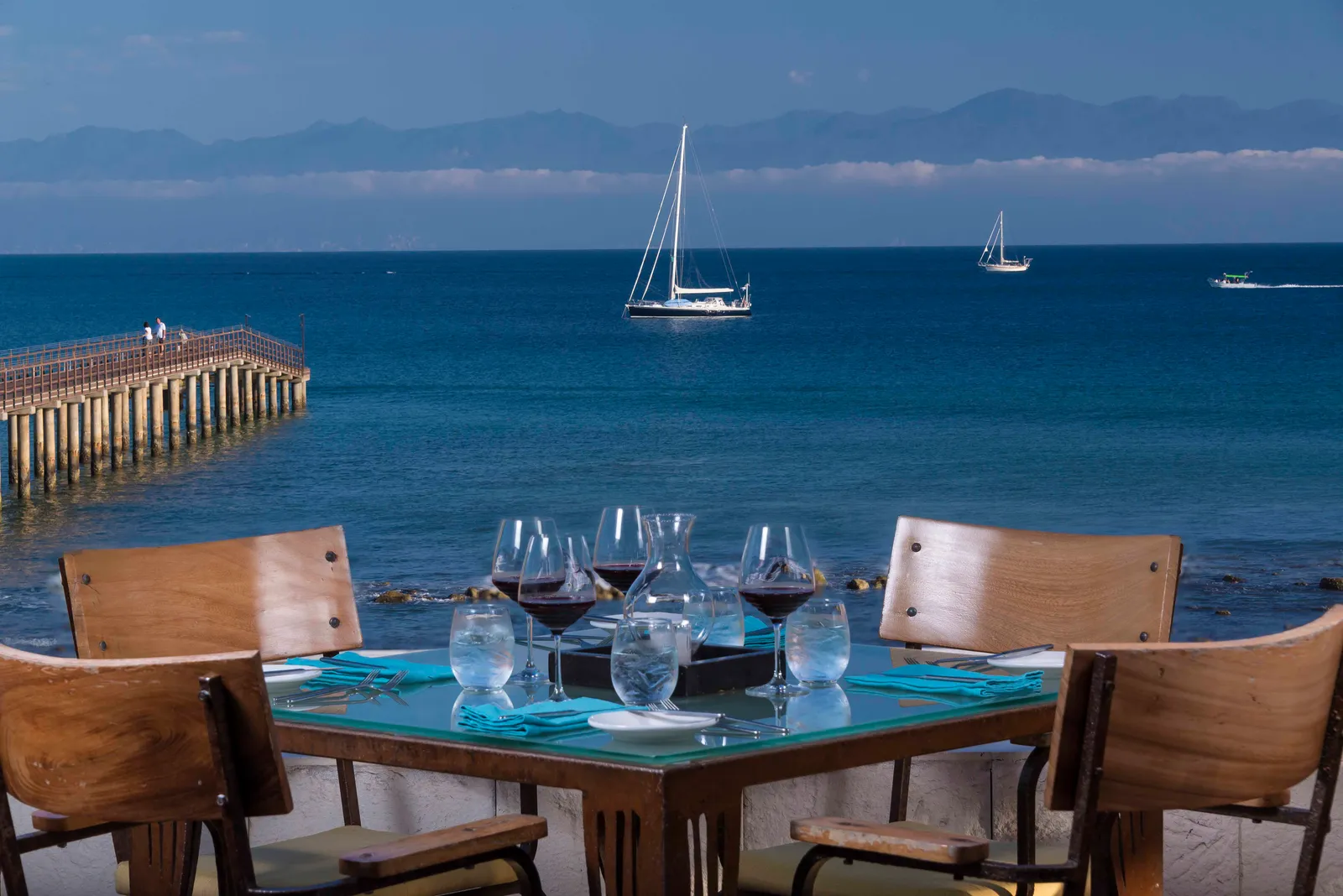 A table set with wine and turquoise linens at the ocean’s edge, sailboats on Banderas Bay and the mountains beyond