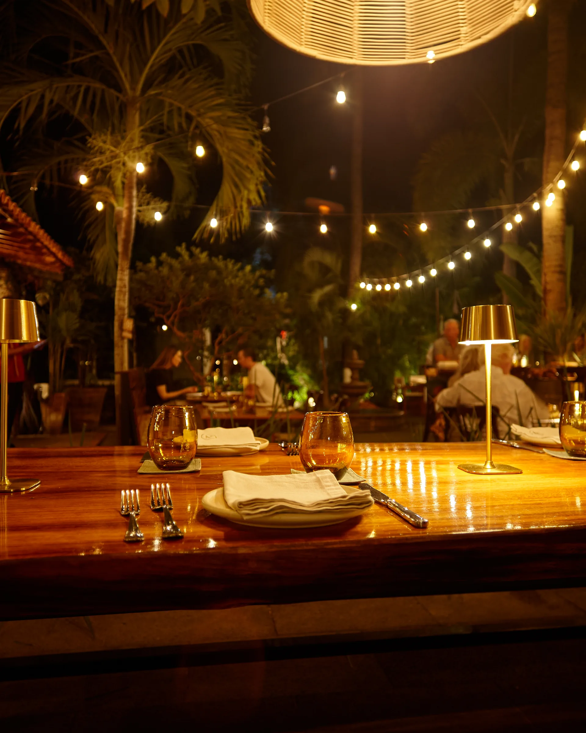 NAEF’s garden at night — a long polished-wood table under string lights, lamps glowing among the palms