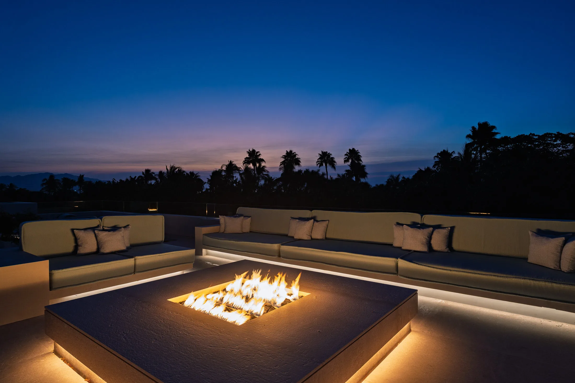 The rooftop fire table lit at dusk, sofas glowing and palms against the last light