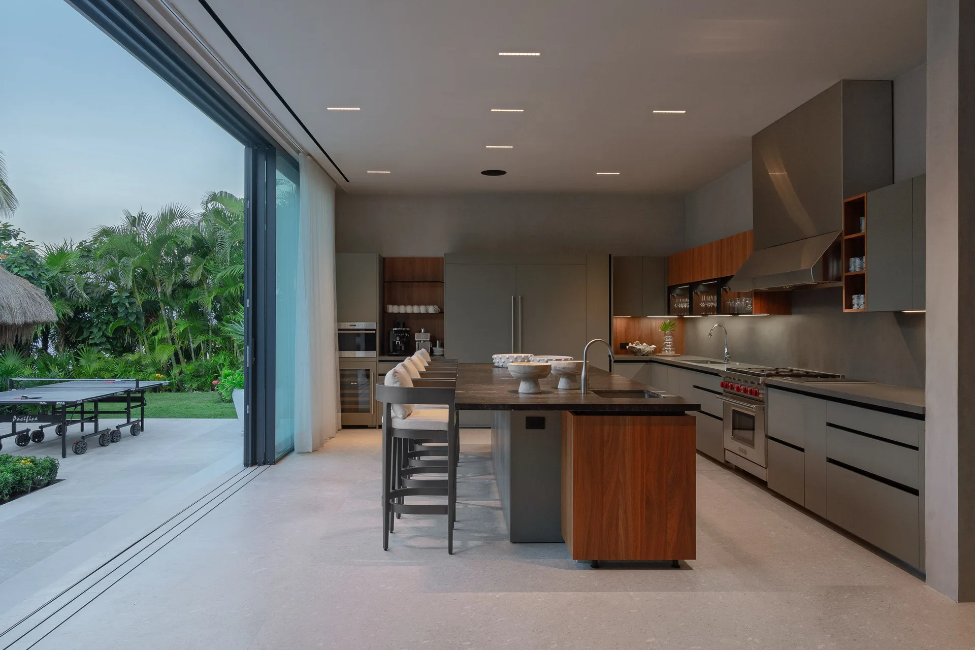 The show kitchen at dusk — stools down the stone island, the Wolf range behind, the ping-pong terrace through the open glass