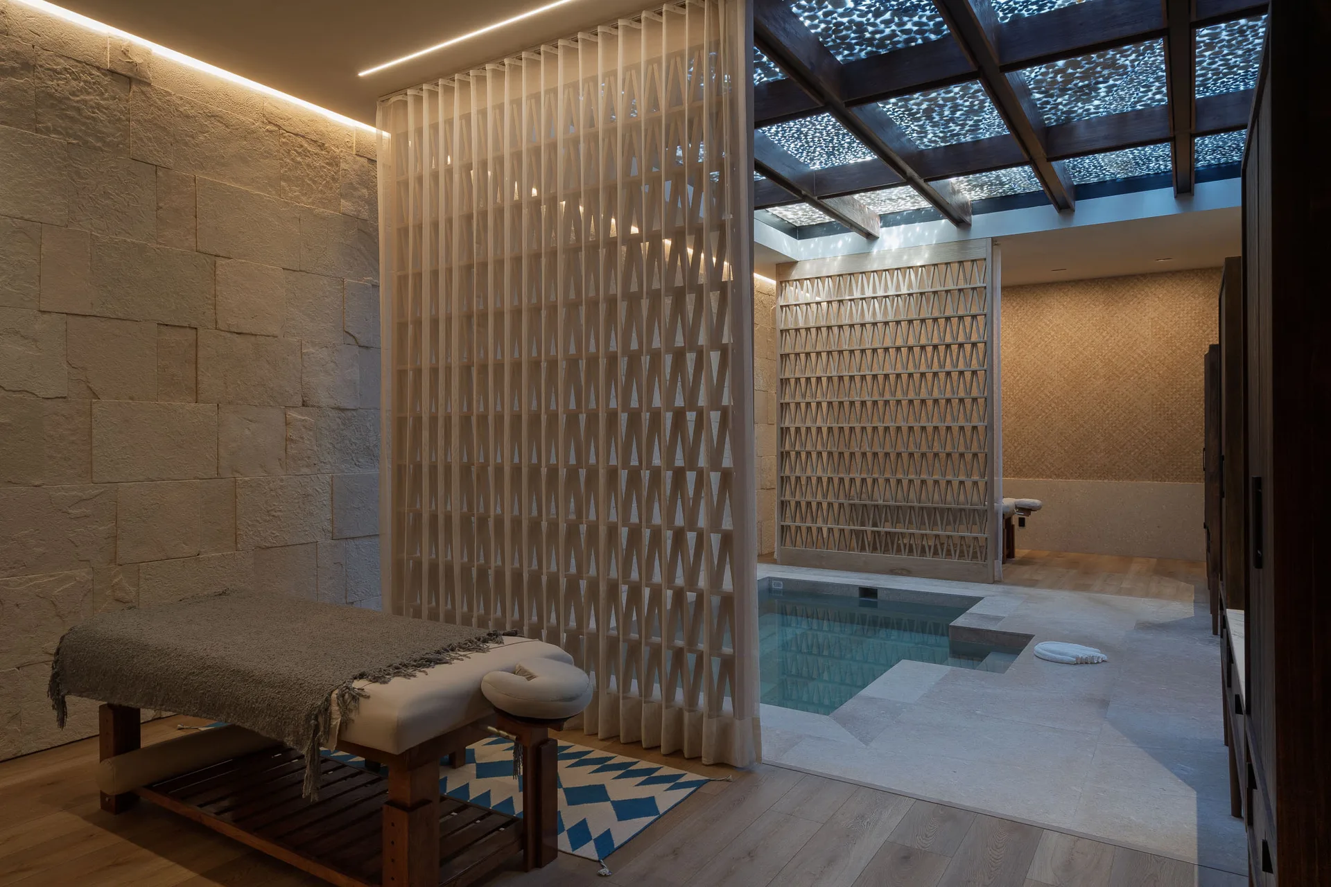 The spa suite — massage table, cold plunge and lattice screens beneath a light-dappled ceiling
