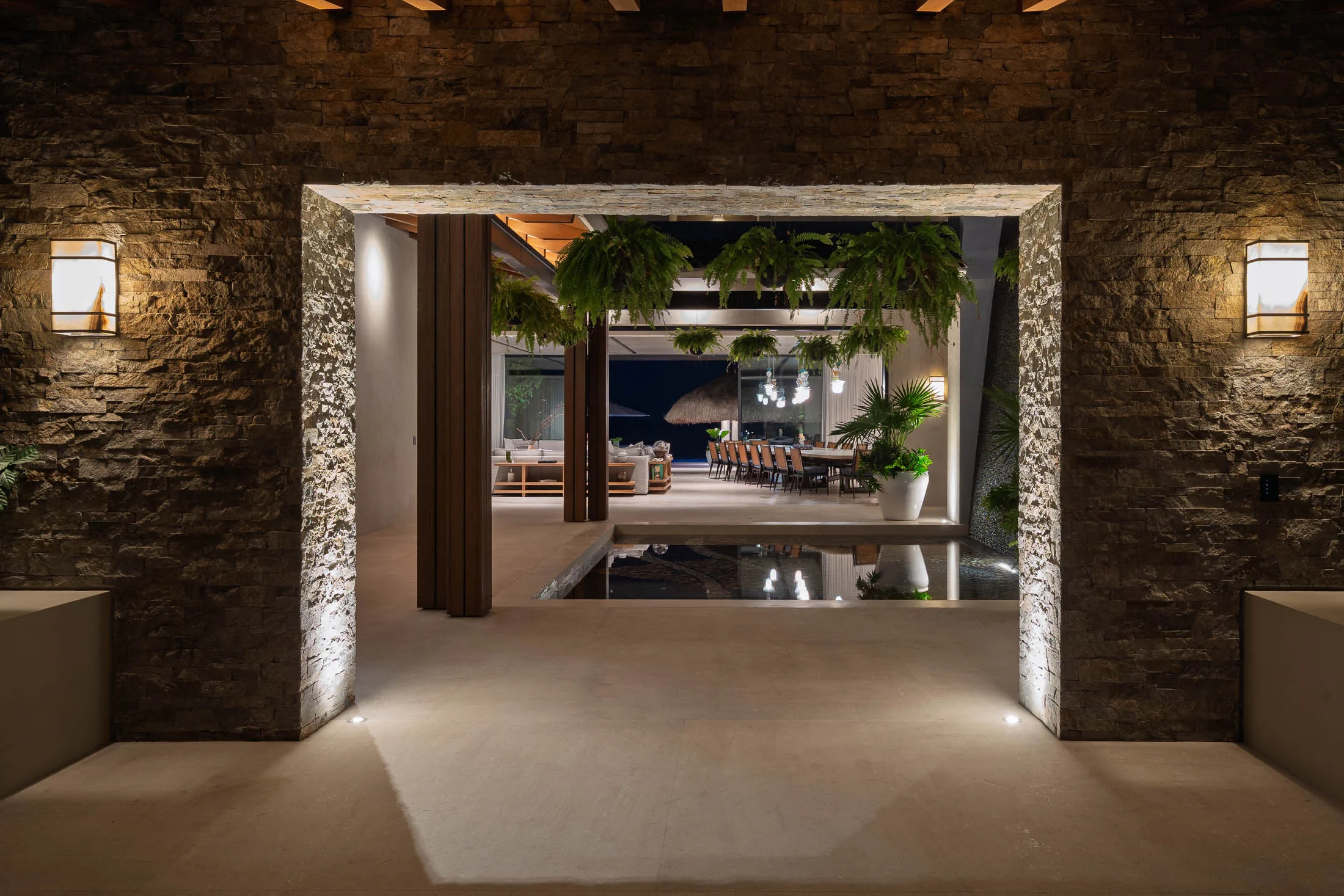 Inside the great room at dusk — ferns overhead, the house framing the Pacific beyond